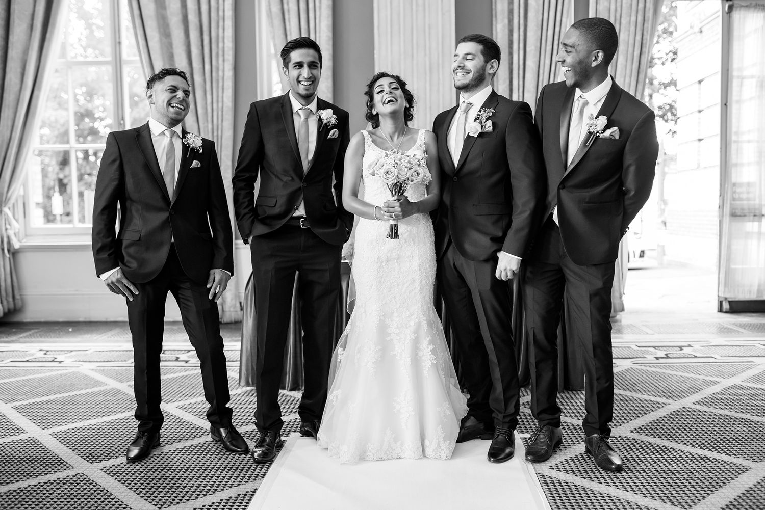 Wedding party walking and laughing together in formal attire down a corridor with elegant columns.