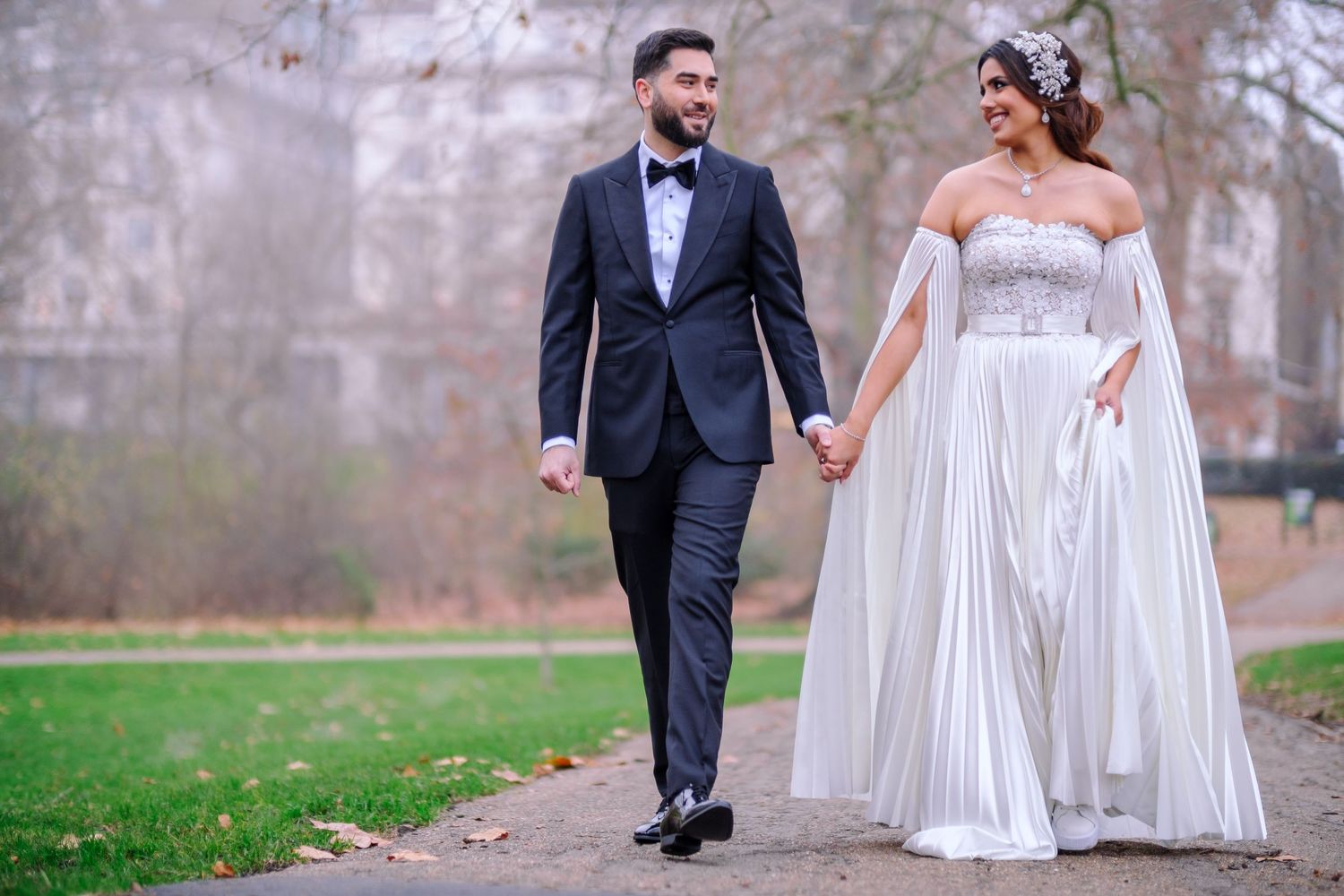 A wedding couple walks hand in hand through a misty park in formal attire during an autumn day.
