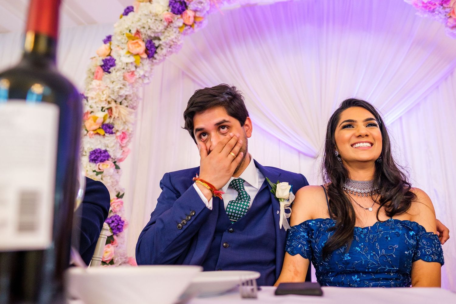 Couple reacting emotionally during wedding reception speeches at a table with floral decorations.