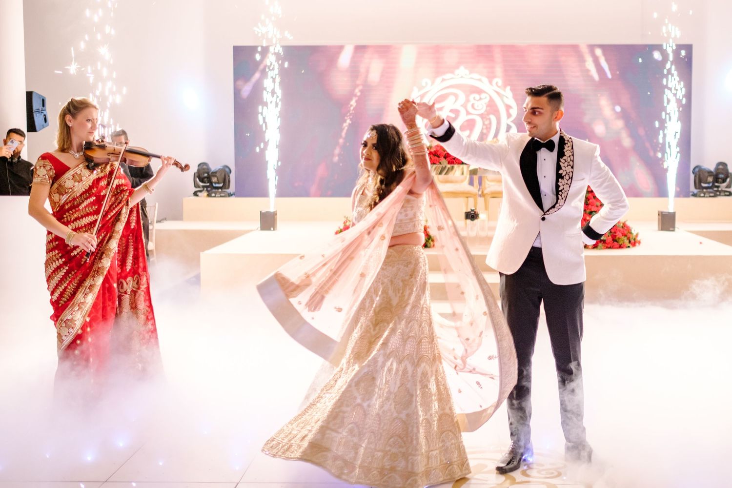 Dancers in traditional Indian wedding attire perform graceful movements on a white illuminated dance floor.
