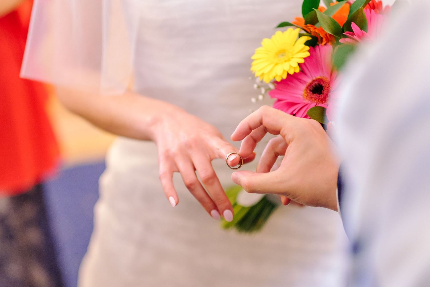 Close-up of hands exchanging wedding rings with colorful gerbera daisies in the background.