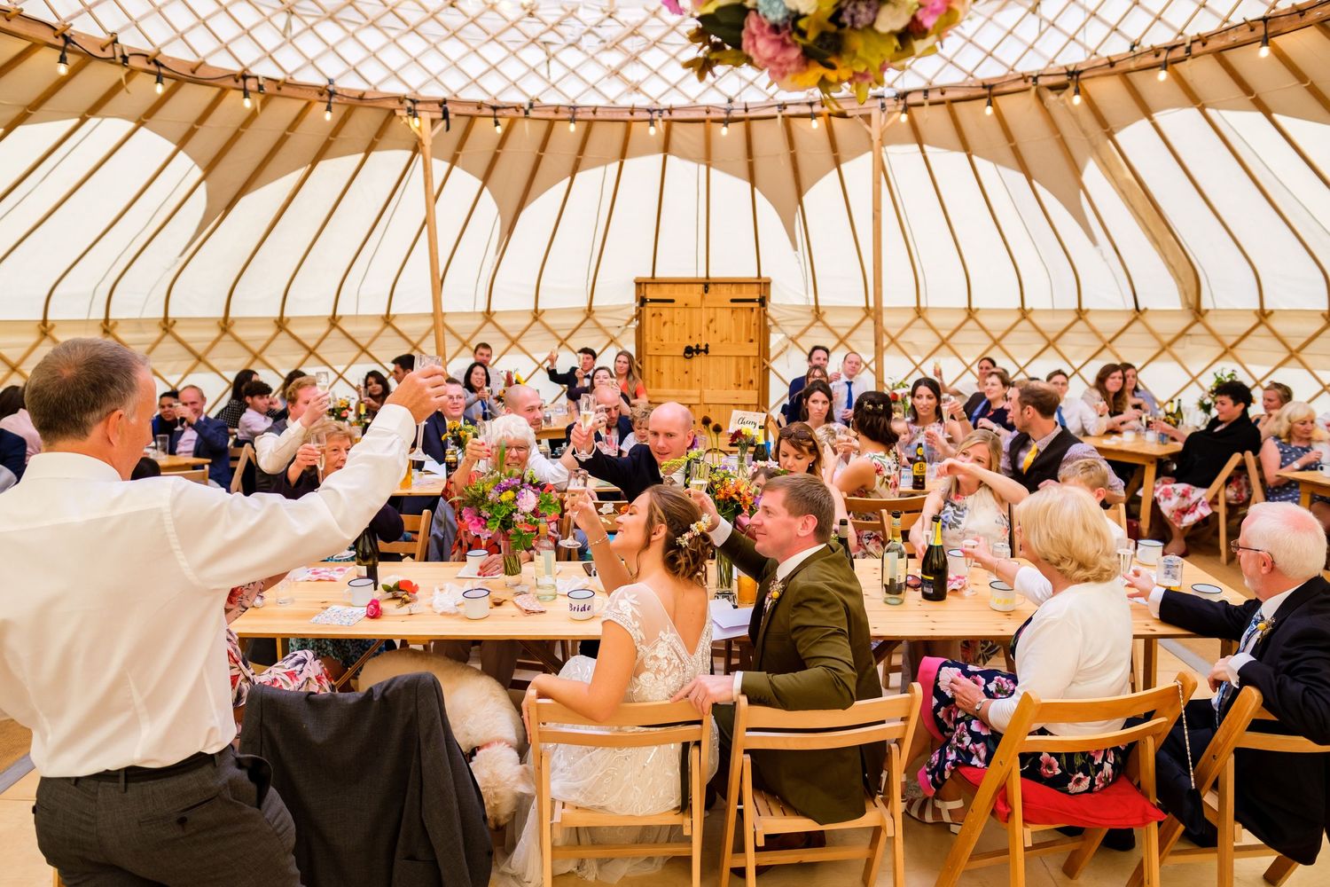 Wedding reception inside a large yurt with wooden tables and decorative lighting.