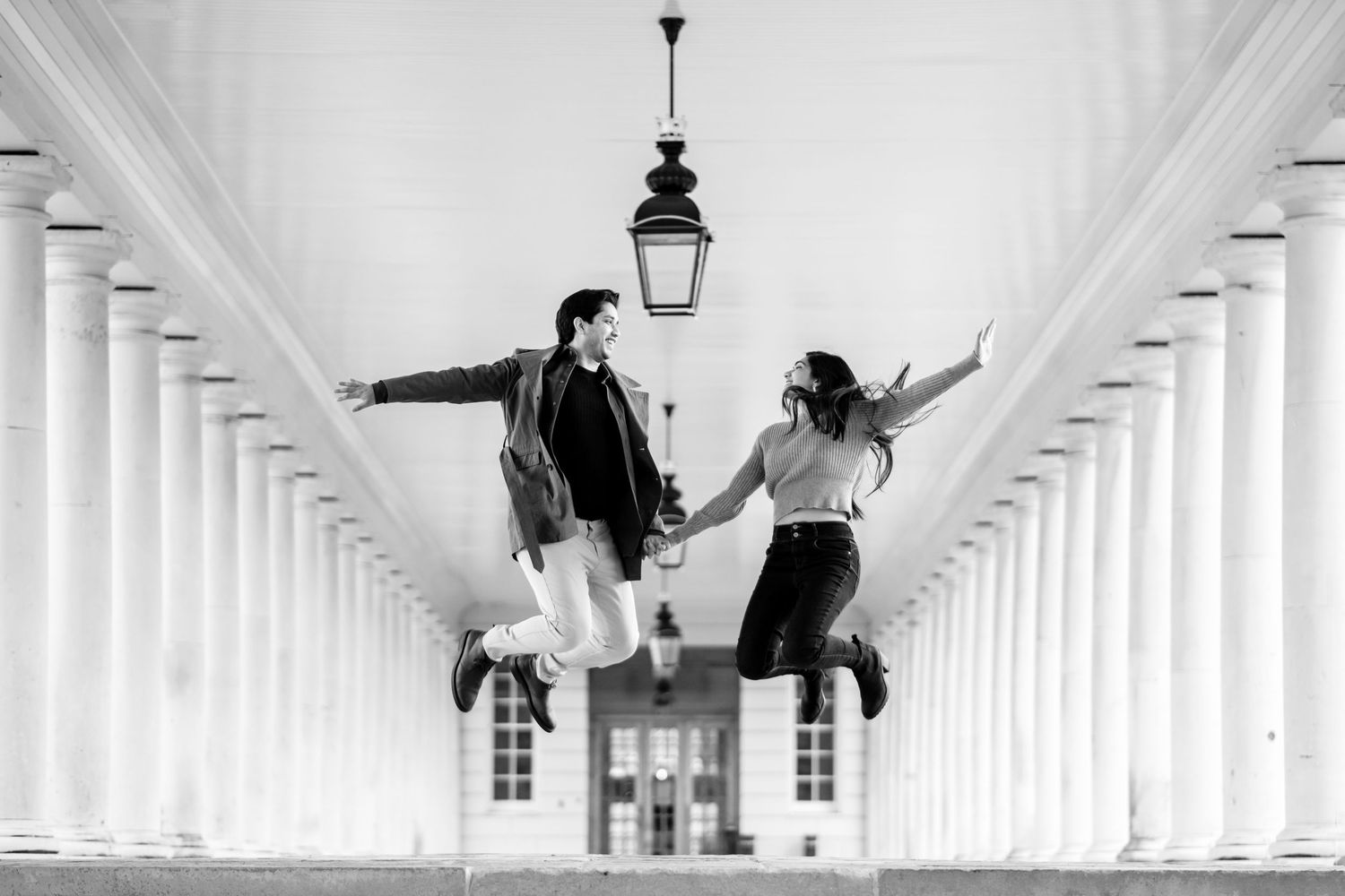 Two people perform gravity-defying parkour moves in a white architectural corridor with repeating columns and arches.