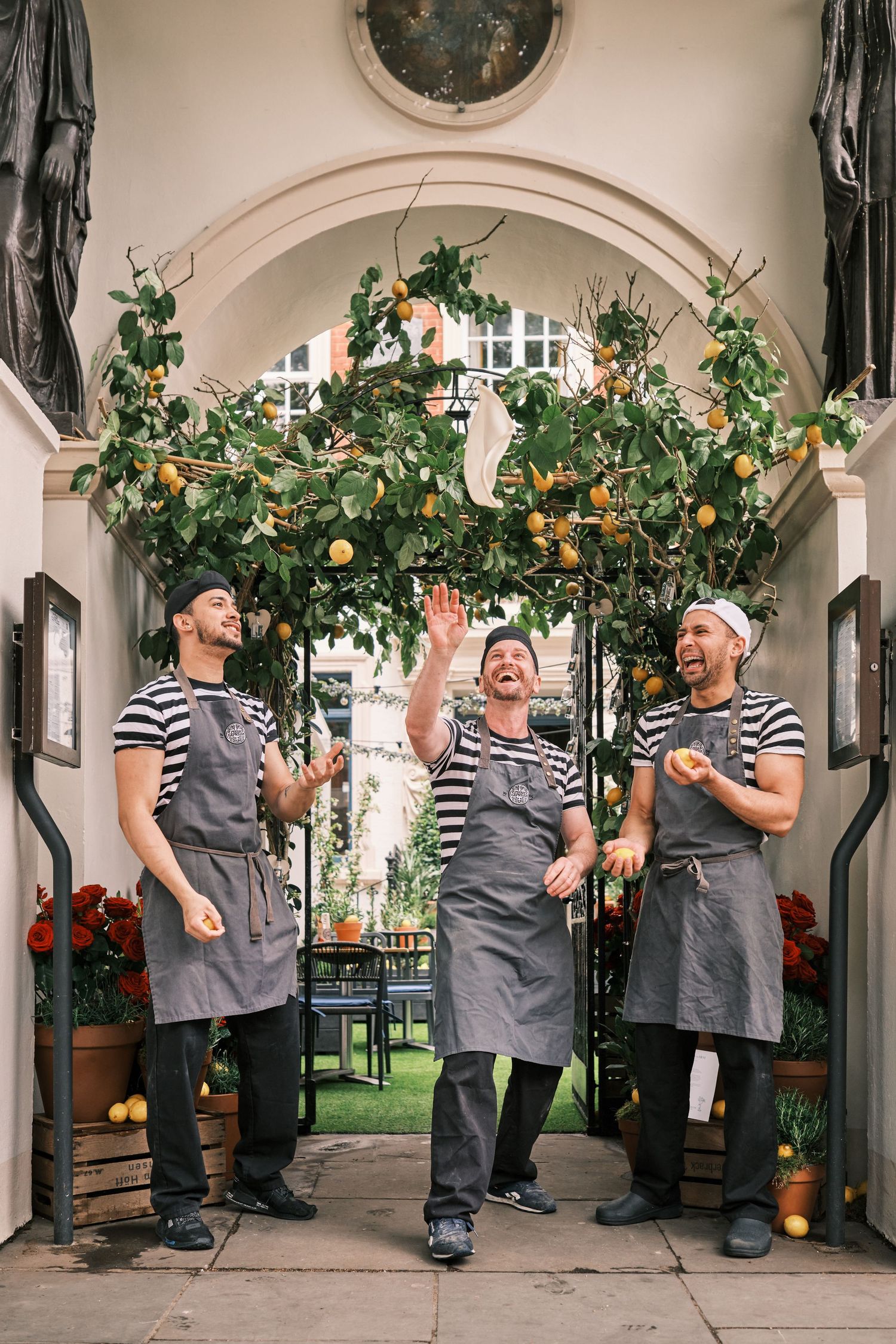 Restaurant staff in striped shirts and gray aprons pose playfully in an outdoor courtyard decorated with hanging plants.