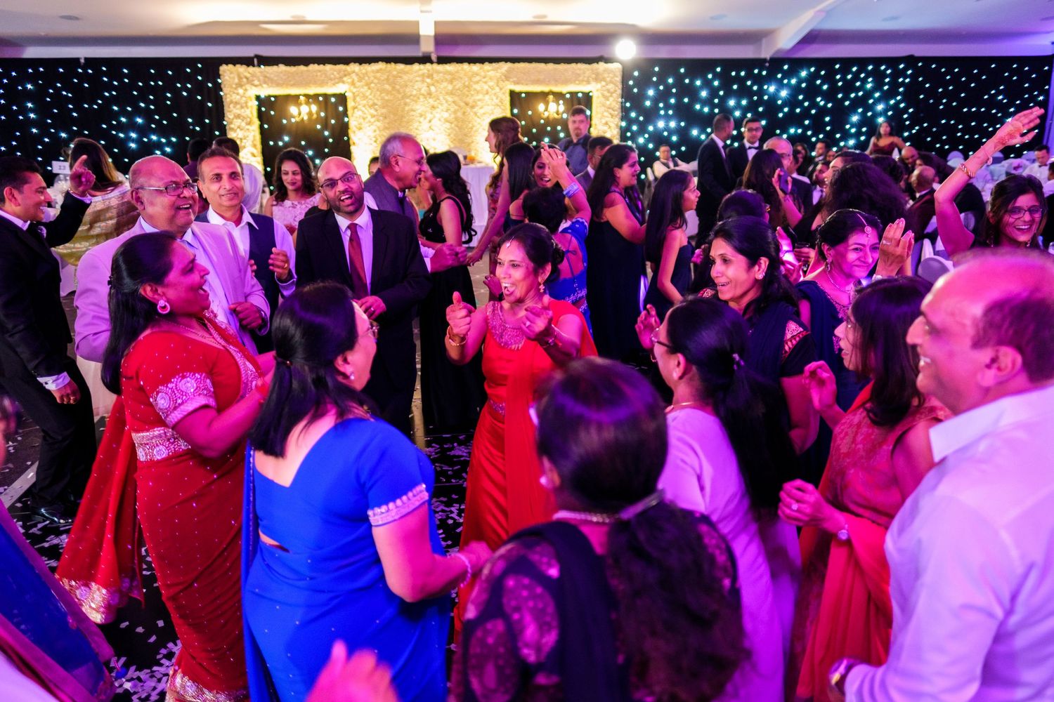 Large crowd dancing and celebrating at a wedding reception with pink and purple mood lighting.