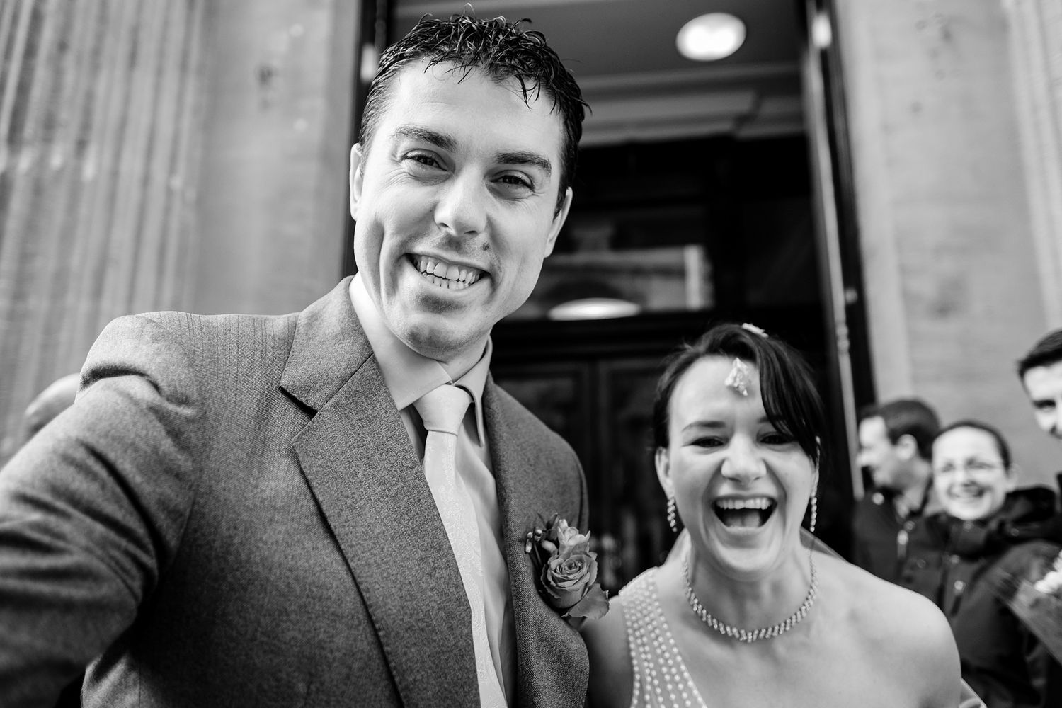 Black and white photo of newlyweds sharing a joyful moment outside a building.