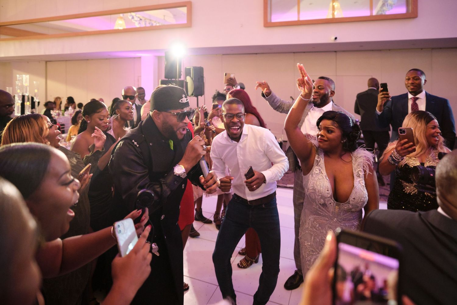 Energetic crowd dancing and celebrating at a wedding reception under pink mood lighting.