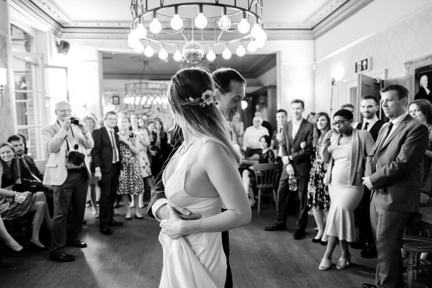 Newlyweds dance together surrounded by wedding guests in elegant venue with chandelier.