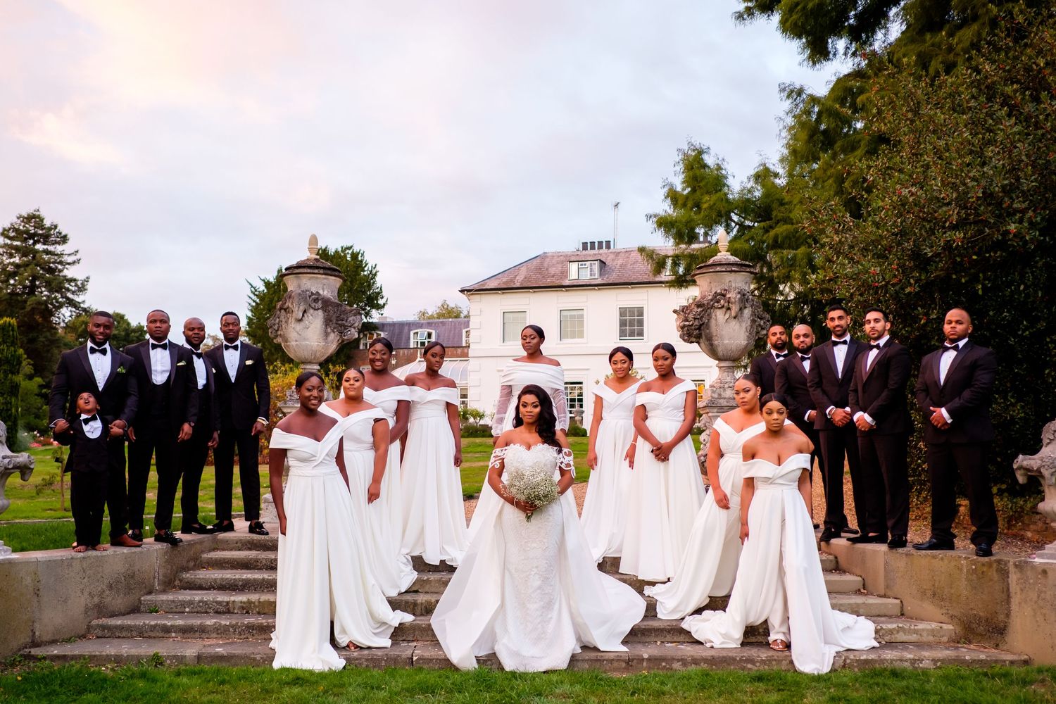 A wedding party poses on grand outdoor stone steps surrounded by trees.
