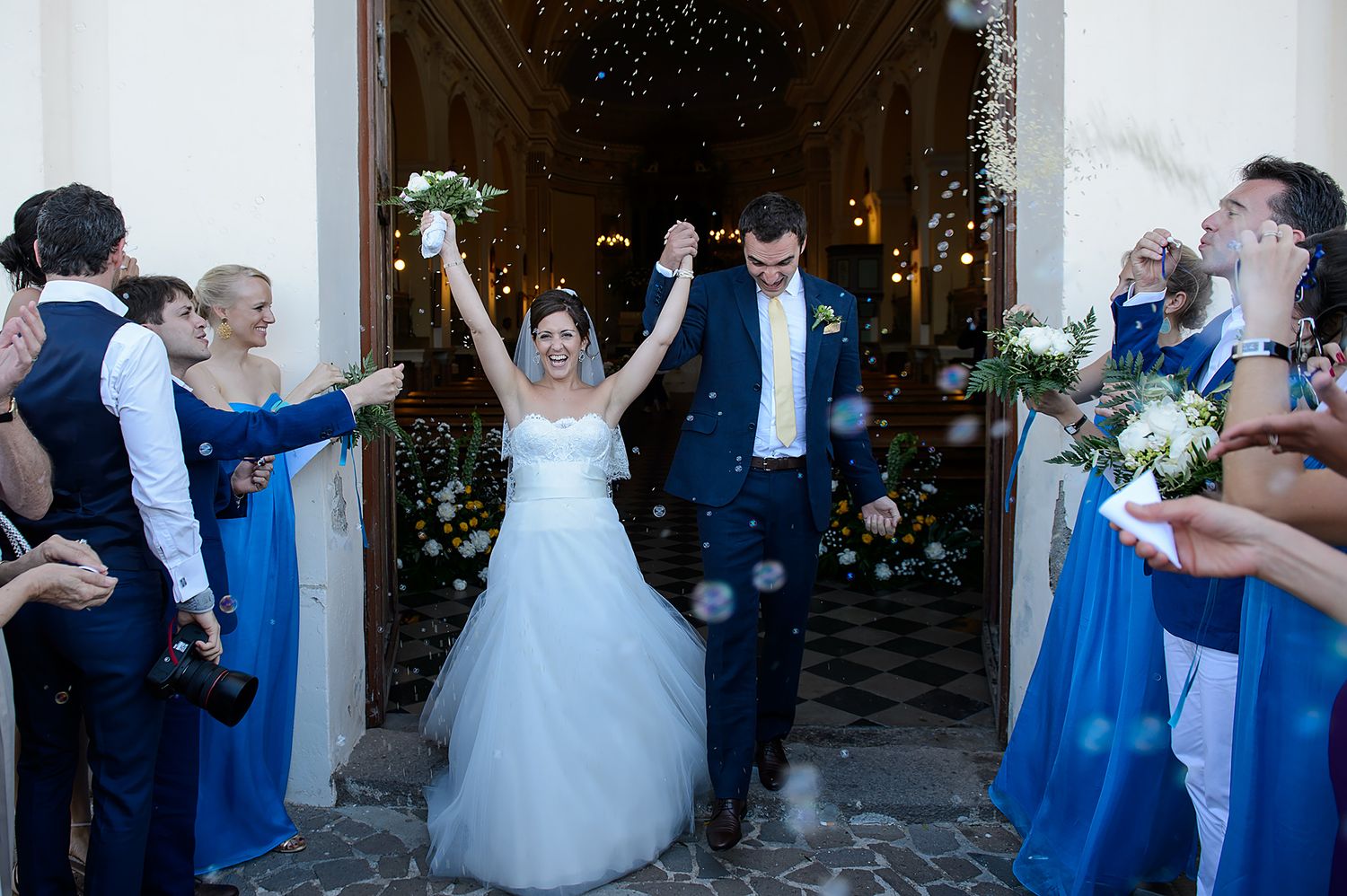 Wedding couple and guests celebrate with dance moves in an elegant reception hall decorated with blue accents and white flowers.