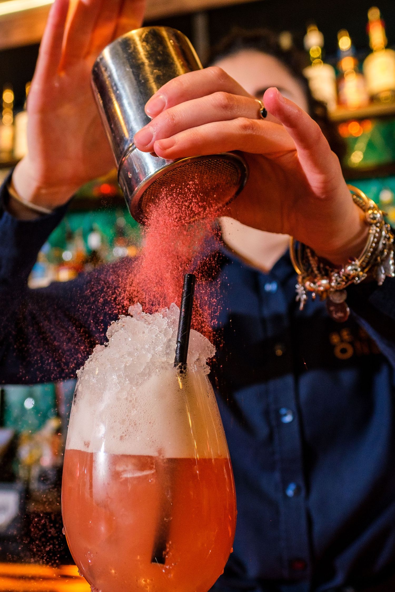 Bartender shaking cocktail with red powder garnish in dim bar lighting while wearing bracelets and dark shirt.