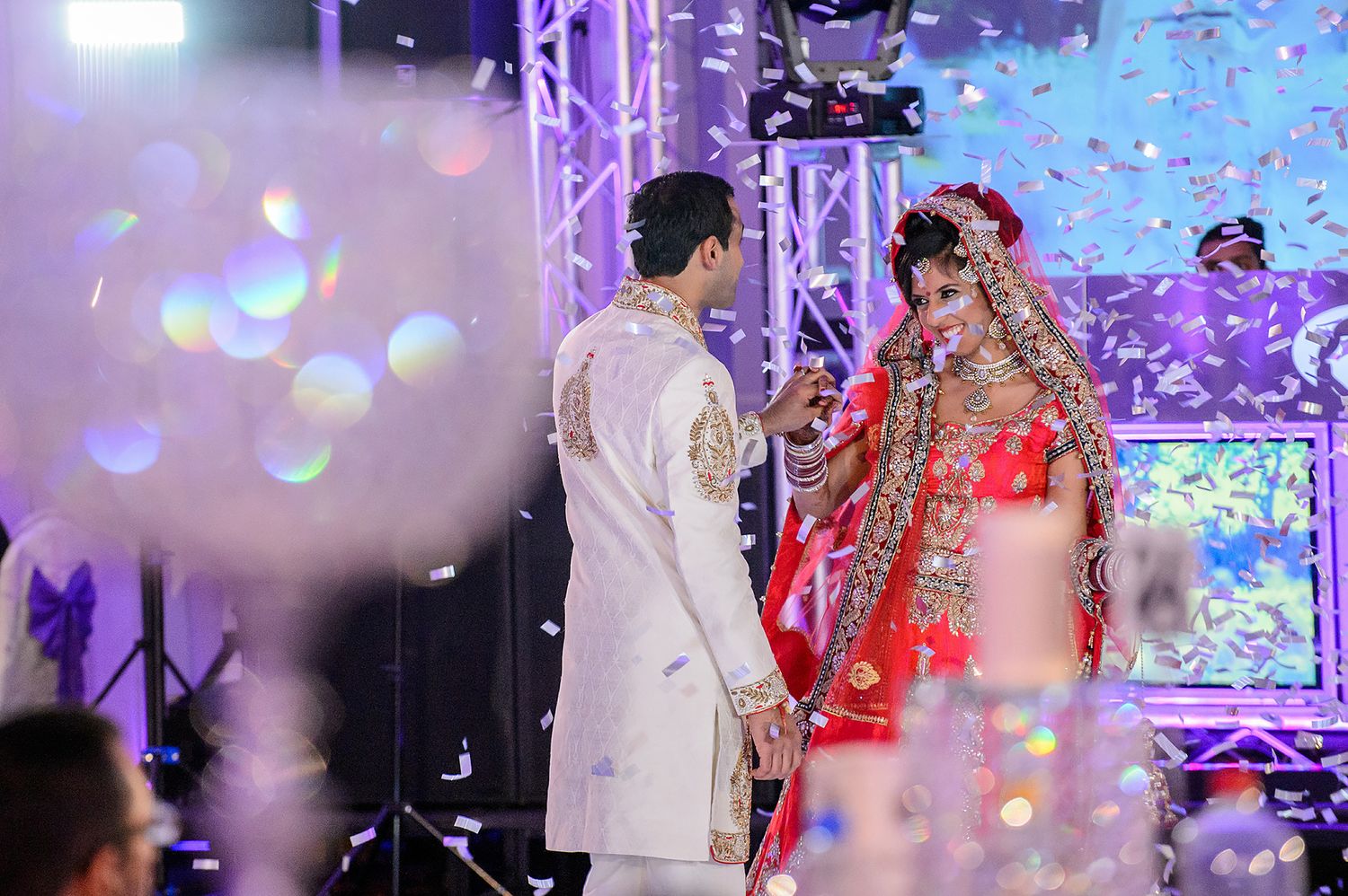 Couple in traditional South Asian wedding attire shares a romantic moment under sparkling lights and confetti.