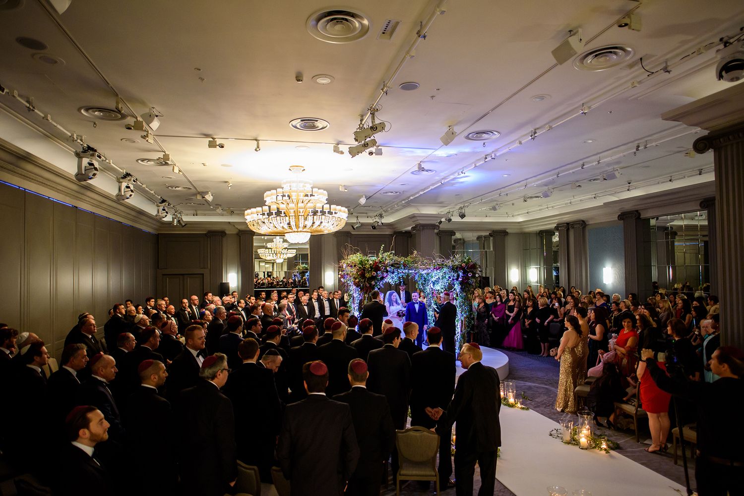 Crowded wedding ceremony in an elegant ballroom with chandelier and dramatic lighting.