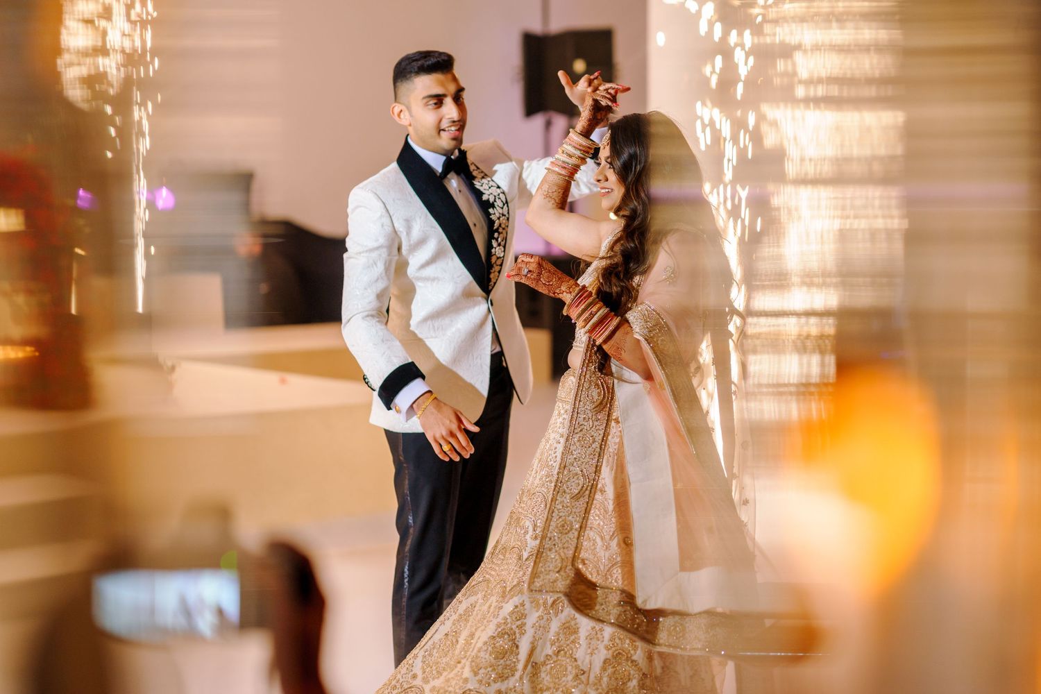 Elegant couple in fusion wedding attire pose against blurred lights and chandelier background.