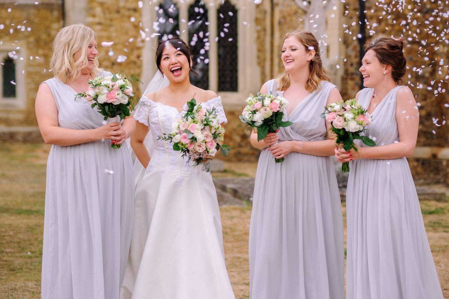 Wedding party wearing lavender bridesmaid dresses laughs together outside stone building during celebration.