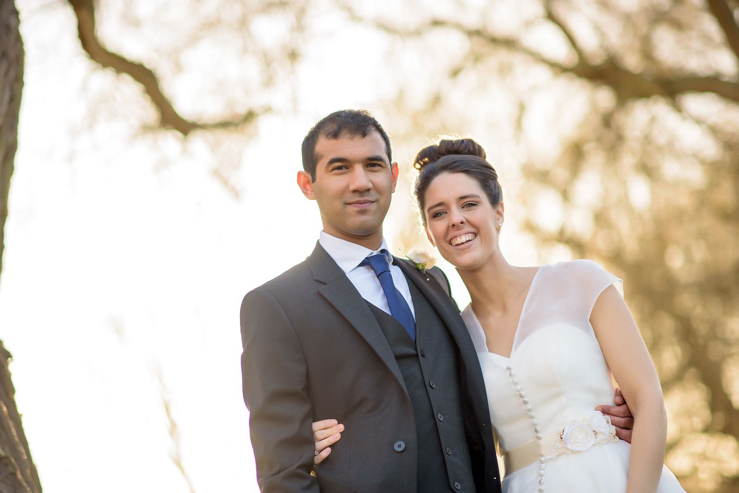 Newlyweds pose together outdoors under winter tree branches in natural light.