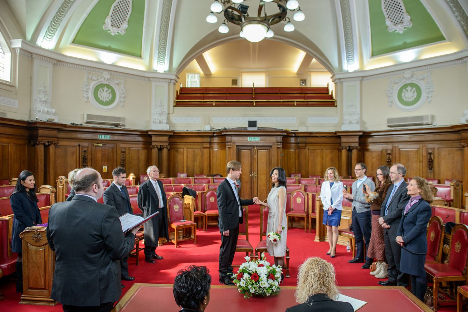 Guests seated in elegant wood-paneled ceremony room with red carpet and ornate chandelier.