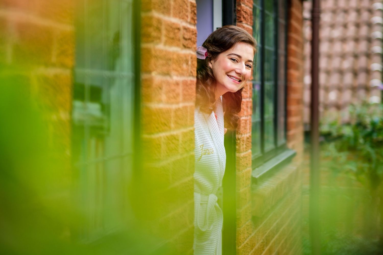 Romantic bridal portrait through greenery with brick wall and window background creating dreamy ambiance.