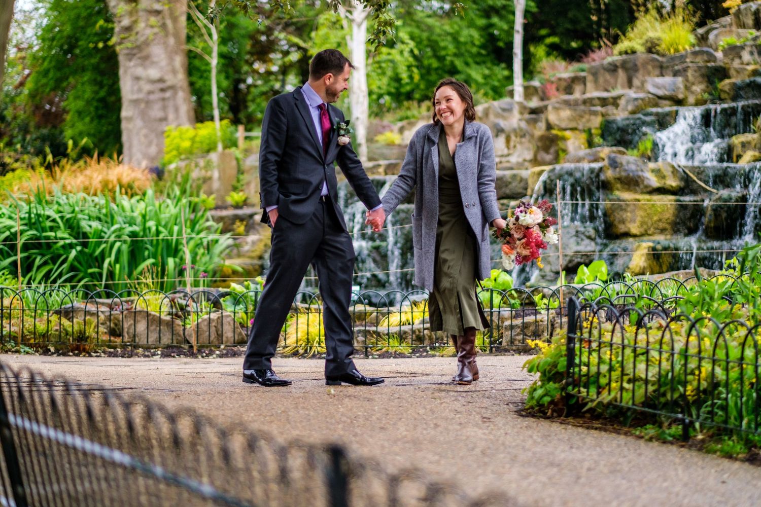 People walk along a lush garden path with stone walls and flowering plants on a sunny day.