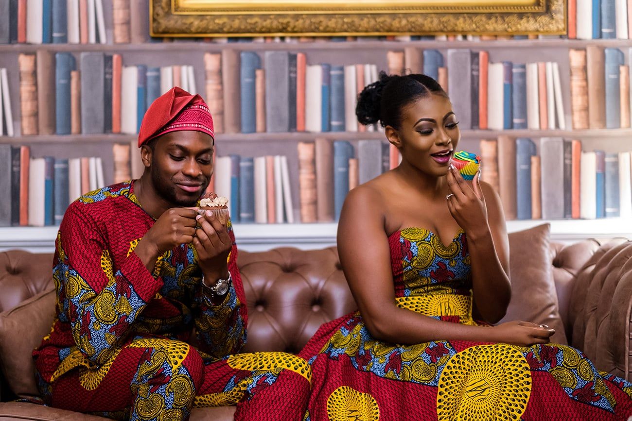 Group wearing colorful African print attire lounges on brown leather sofa in library setting.