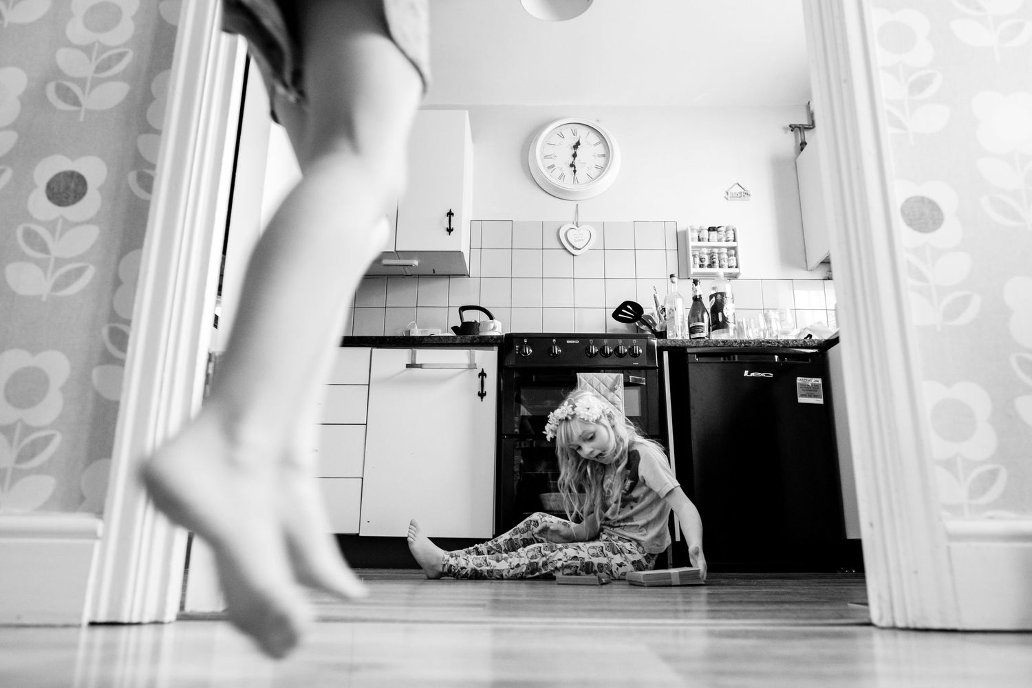 Black and white photo of a dog lying on a kitchen floor with someone's feet visible in the doorway.