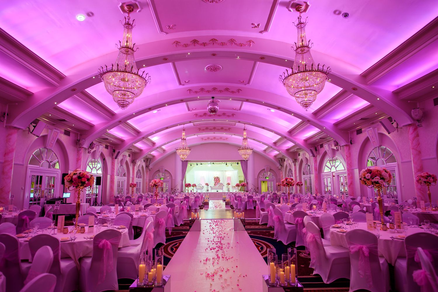 Luxurious pink-lit wedding reception hall with crystal chandeliers and elegant table settings.
