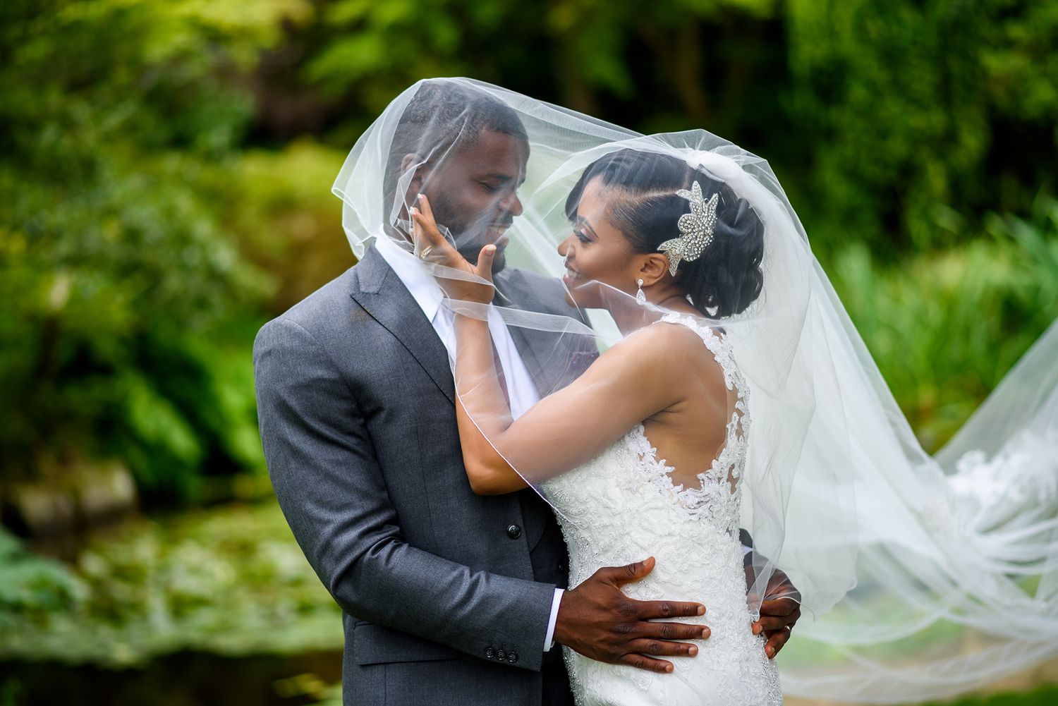 Elegant wedding couple embraces under a flowing veil in a garden setting.