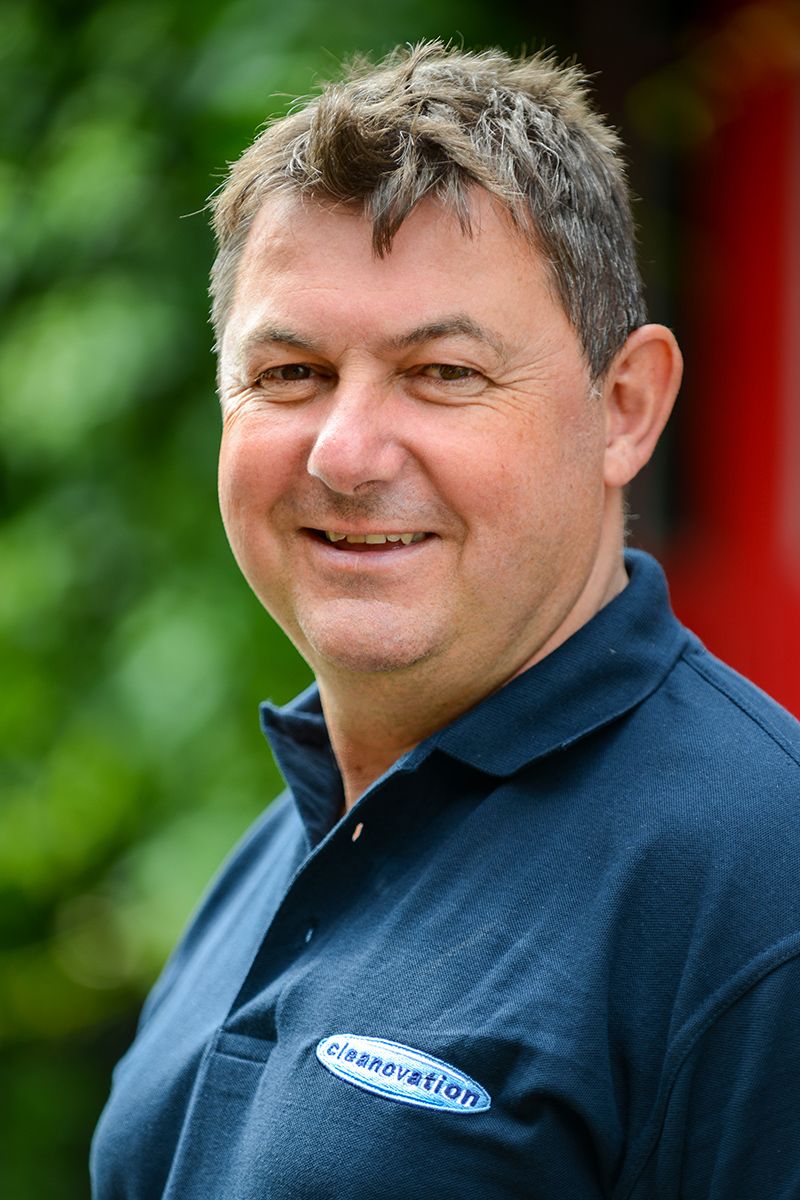 Person wearing navy blue polo shirt with company logo smiles warmly outdoors against green foliage.