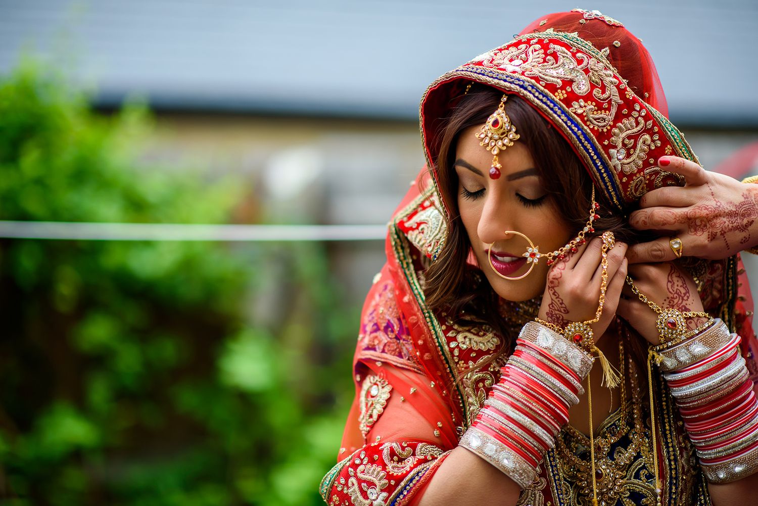 A bride in an ornate red and gold Indian wedding dress adjusts her jewelry and veil.