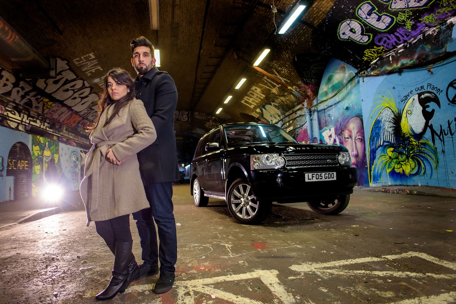 People exploring a graffiti-covered underground area with car headlights visible.