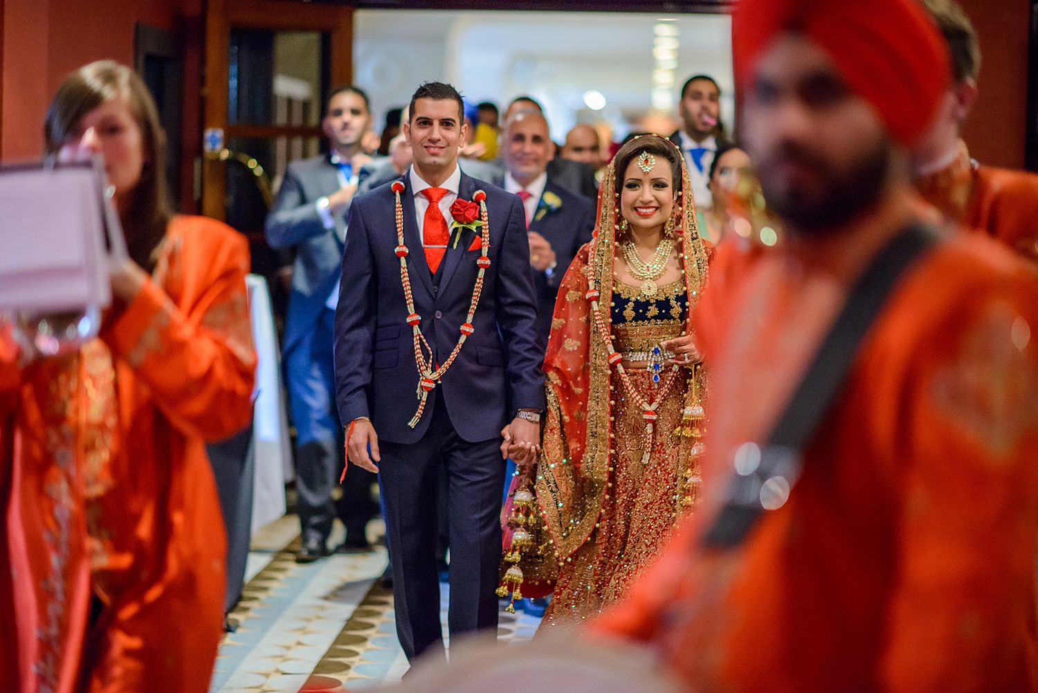 Traditional Indian wedding procession with guests in formal attire and bright orange decorations.