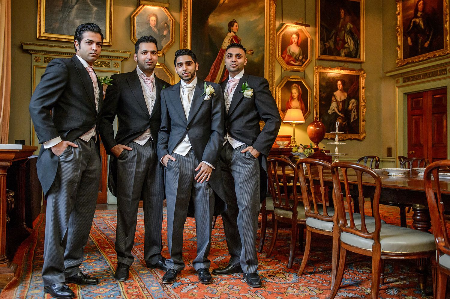 Groomsmen in dark suits pose together in an elegant room with traditional portrait paintings.