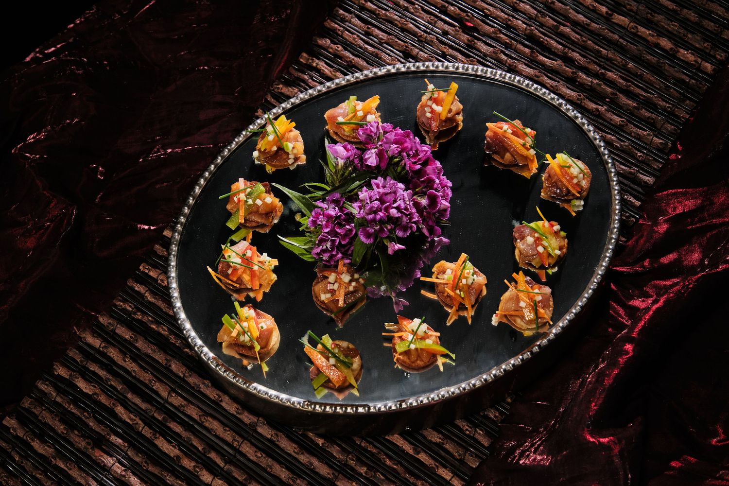 Elegant black platter with decorative bite-sized appetizers arranged around purple flower garnish on dark fabric.