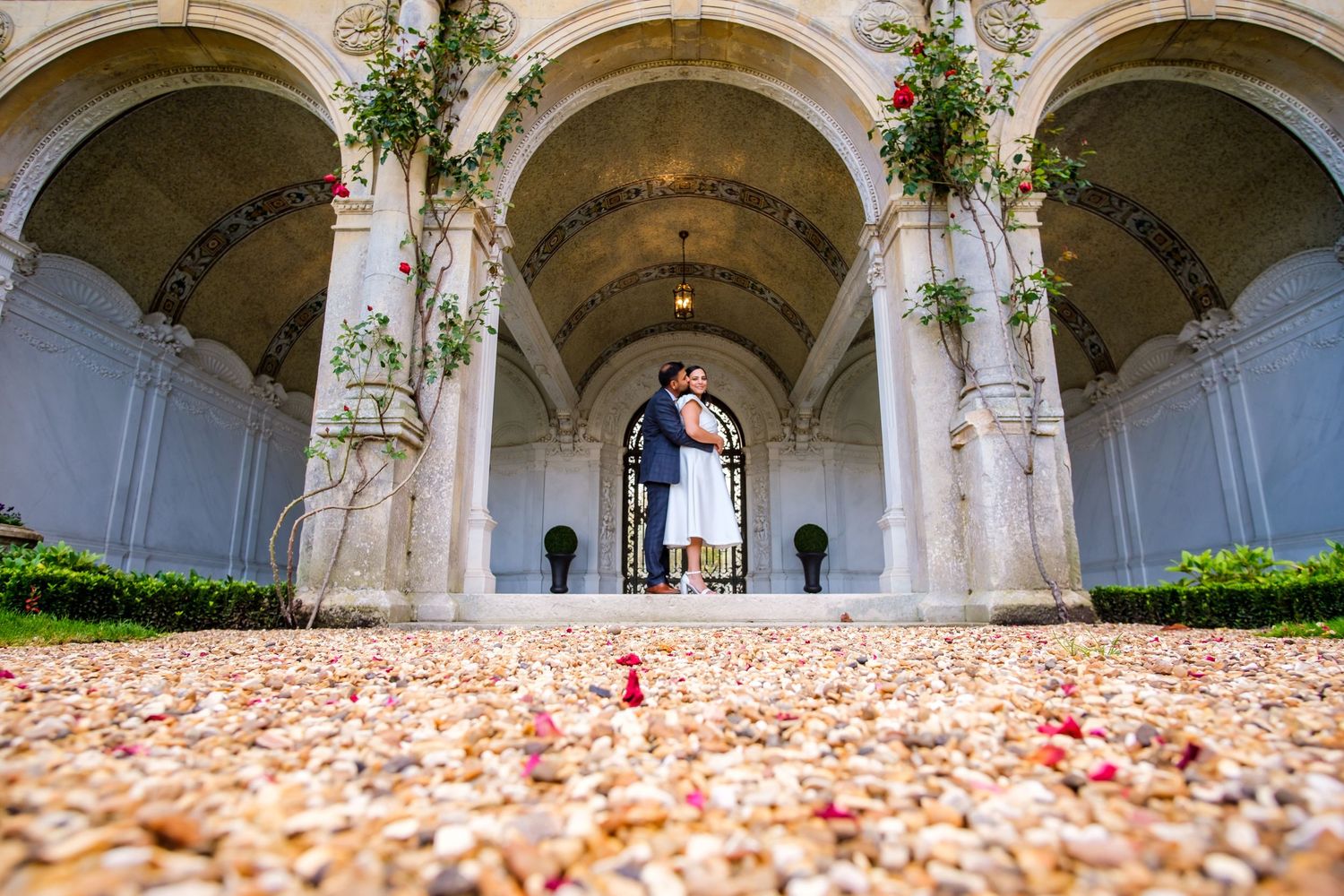 Stone archways with climbing vines line a walkway covered in scattered rose petals creating a romantic atmosphere.