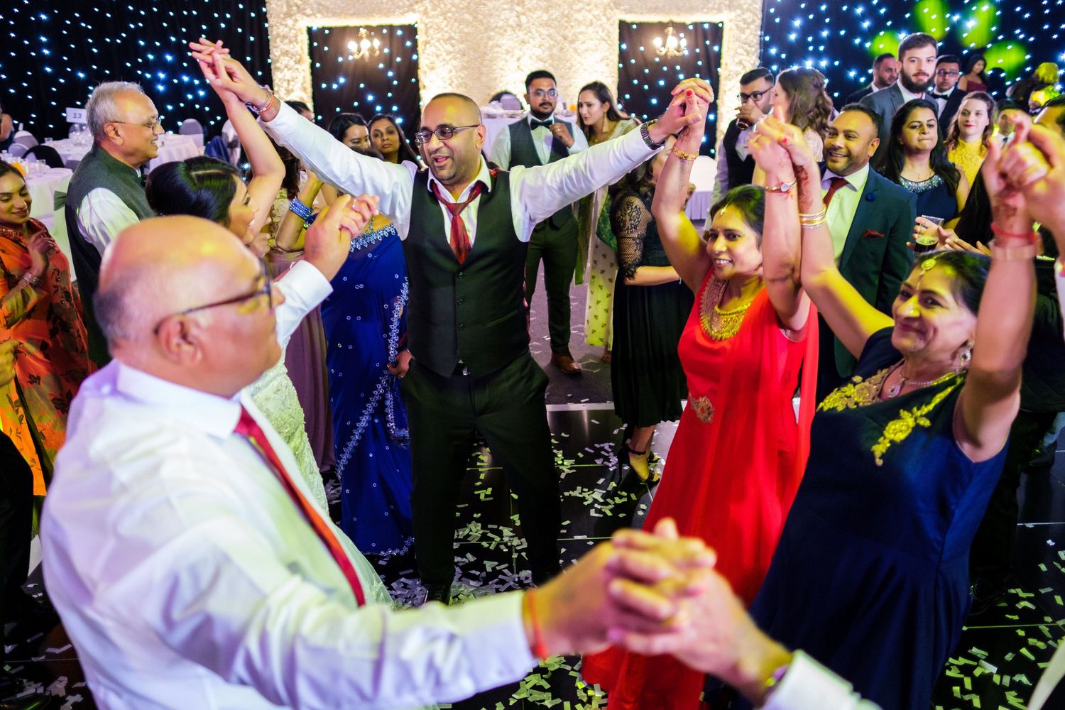 Wedding guests dancing and raising their hands in celebration with confetti and blue lighting effects.