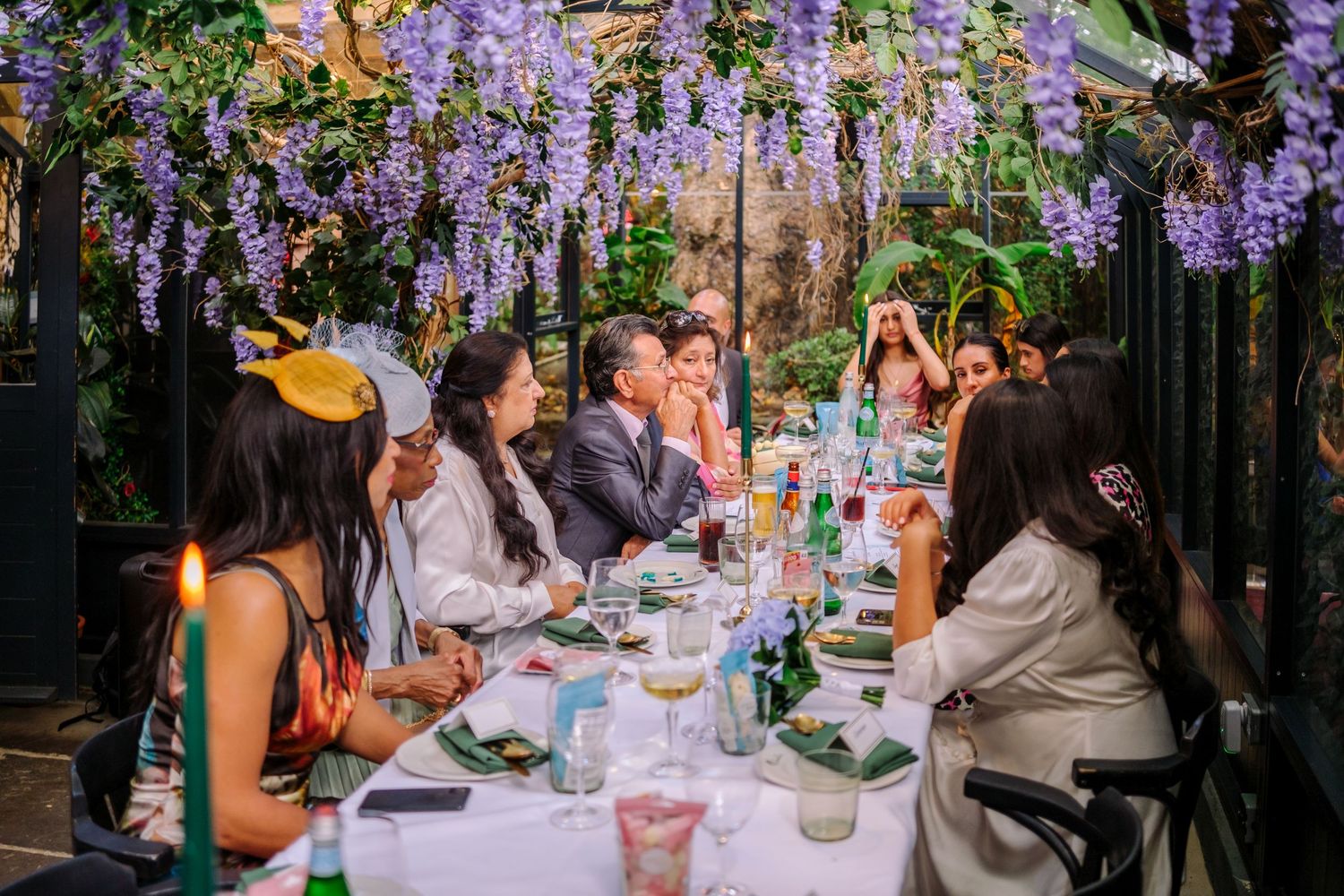 Wedding reception guests dining under hanging purple wisteria in an outdoor garden setting.