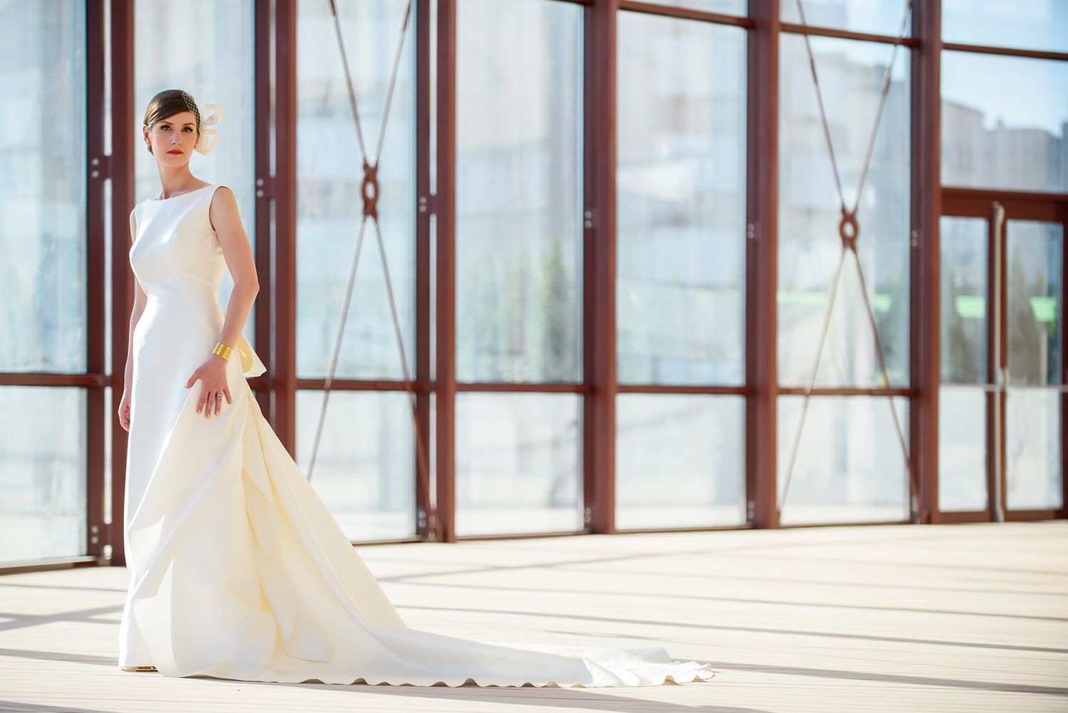 Elegant wedding dress displayed in a bright sunlit glass corridor with reflective floors.
