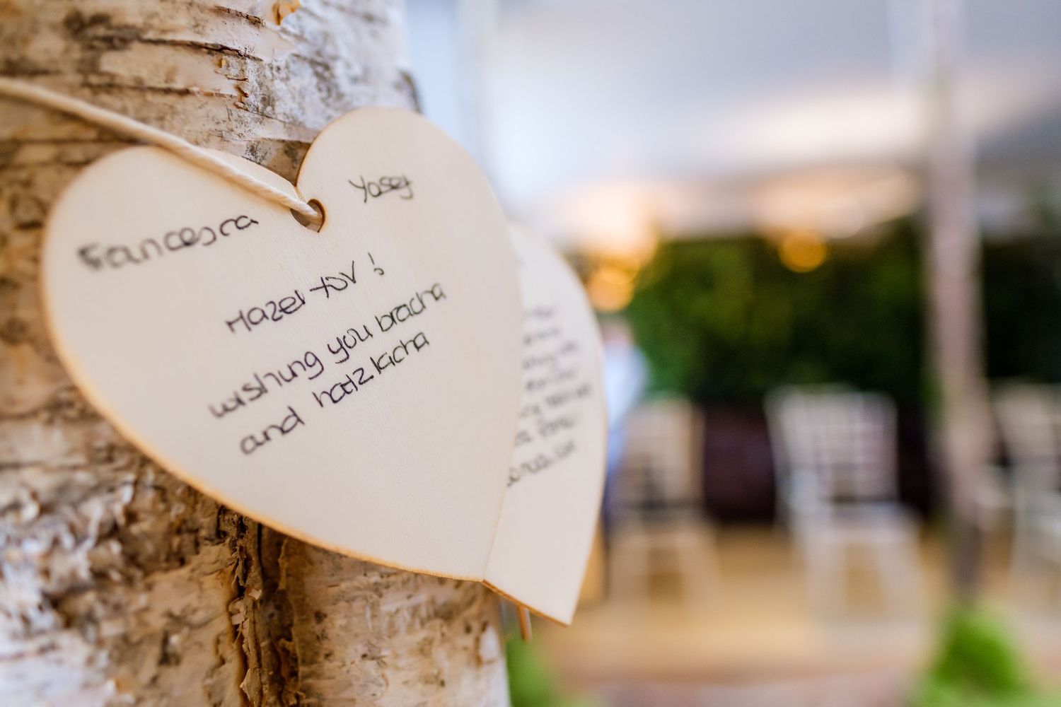 Heart-shaped paper tags hang from rustic tree branches at an outdoor wedding venue with soft lighting.