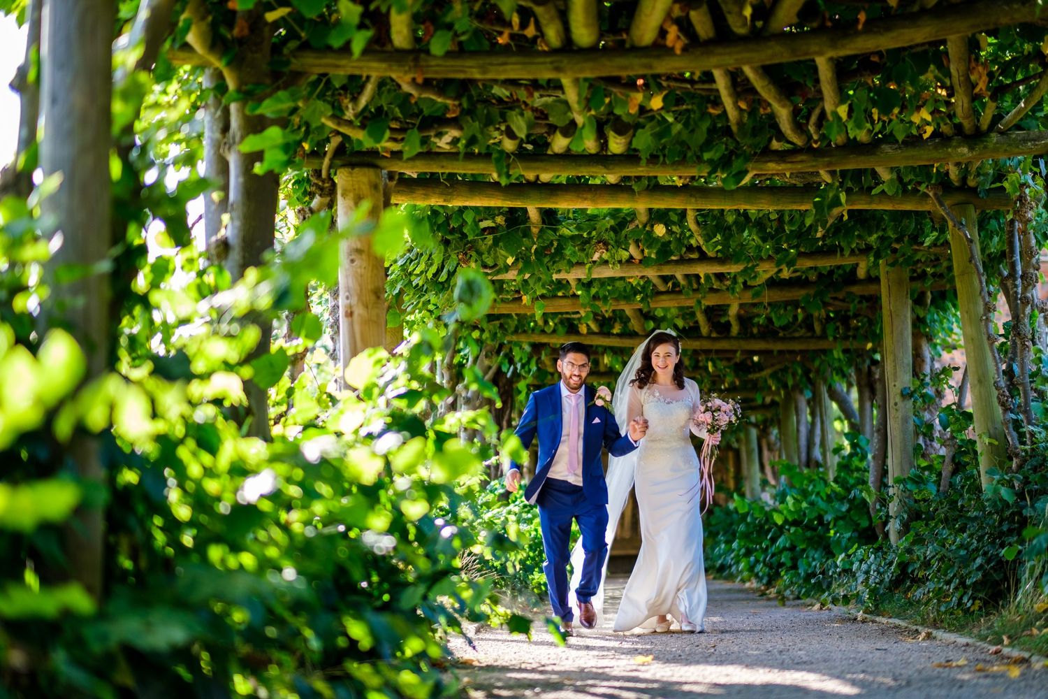 Couple walking through a lush green vine-covered garden pergola.