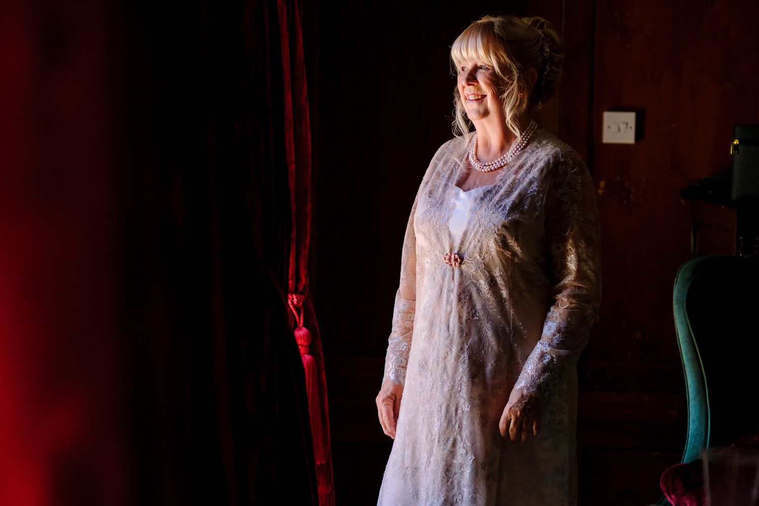 Woman in white lace dress standing against dark red theatrical backdrop with dramatic lighting.