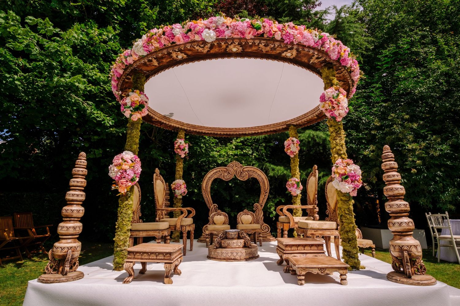 Ornate wedding mandap decorated with pink flowers and gold accents in an outdoor garden setting.