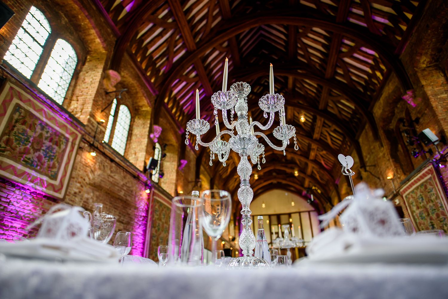 Ornate wooden beam ceiling decorated with chandeliers and purple uplighting for an elegant reception.