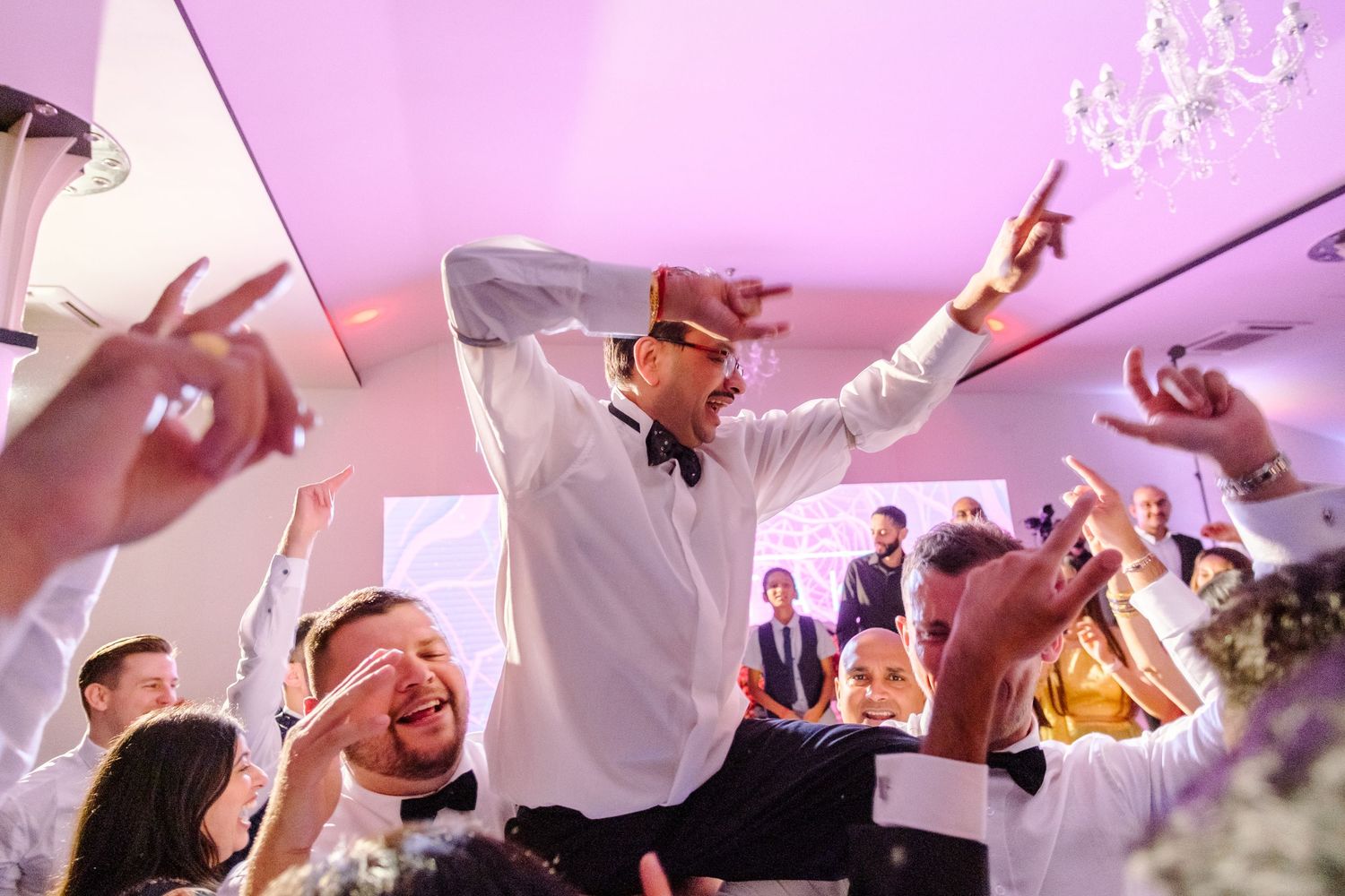 Wedding celebration with guests dancing and raising hands under pink lighting.