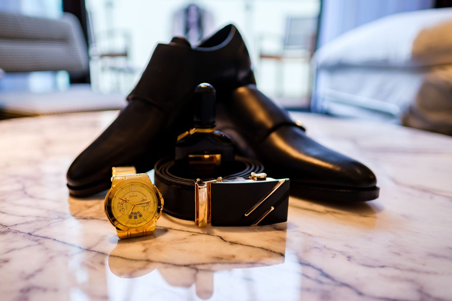 Black leather dress shoes displayed with a gold watch and black leather belt on marble surface.