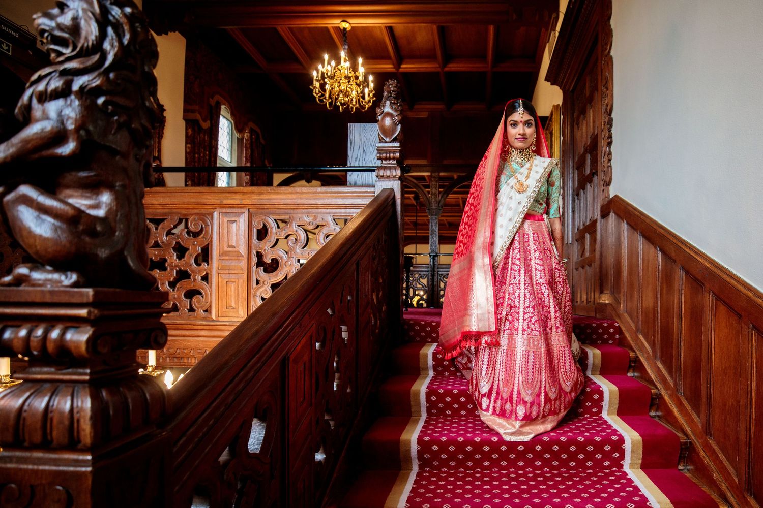 Red and gold traditional wedding attire photographed in an ornate wood-paneled hallway with chandelier.