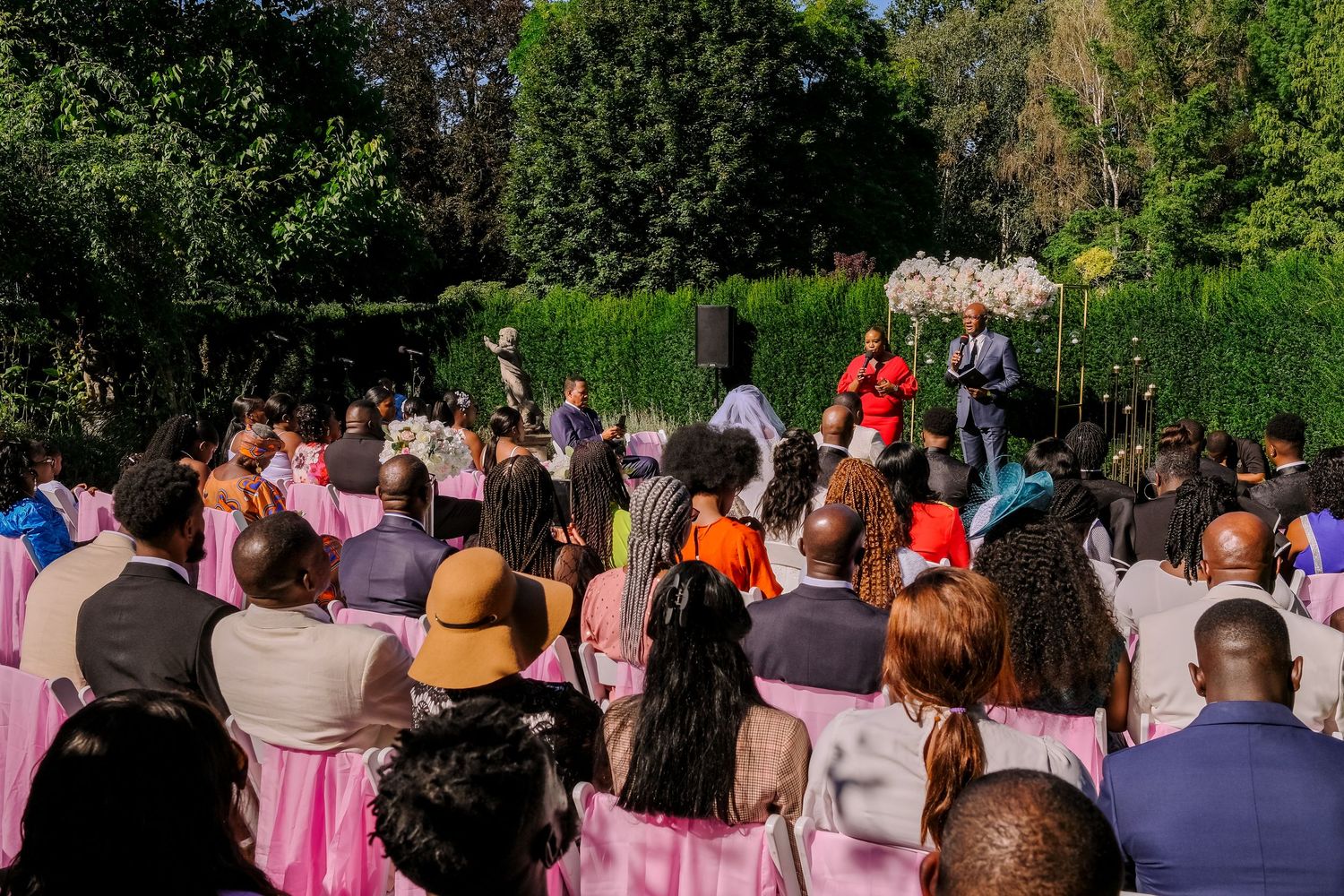 Wedding guests stand in rows outdoors watching a ceremony under green trees on a sunny day.