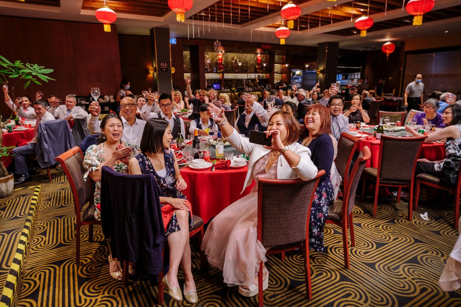 Wedding guests dance and celebrate on a dimly lit ballroom floor with red lantern lighting overhead.