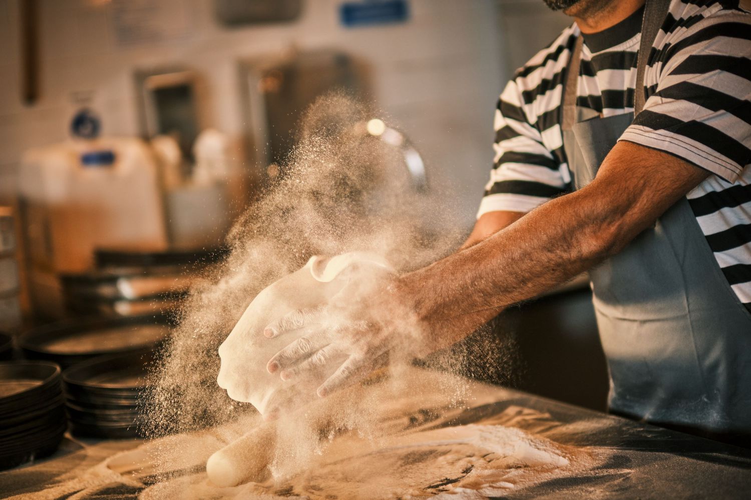 Flour dust billows as hands knead dough on a wooden preparation surface.