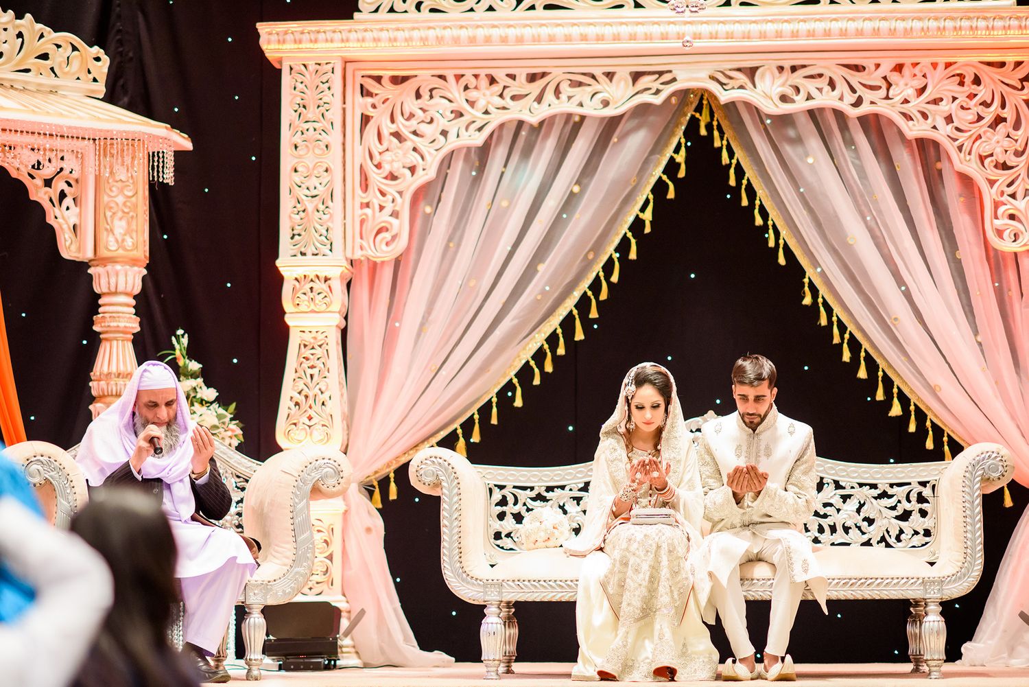 Ornate wedding stage decorated with pink lighting and white drapes.