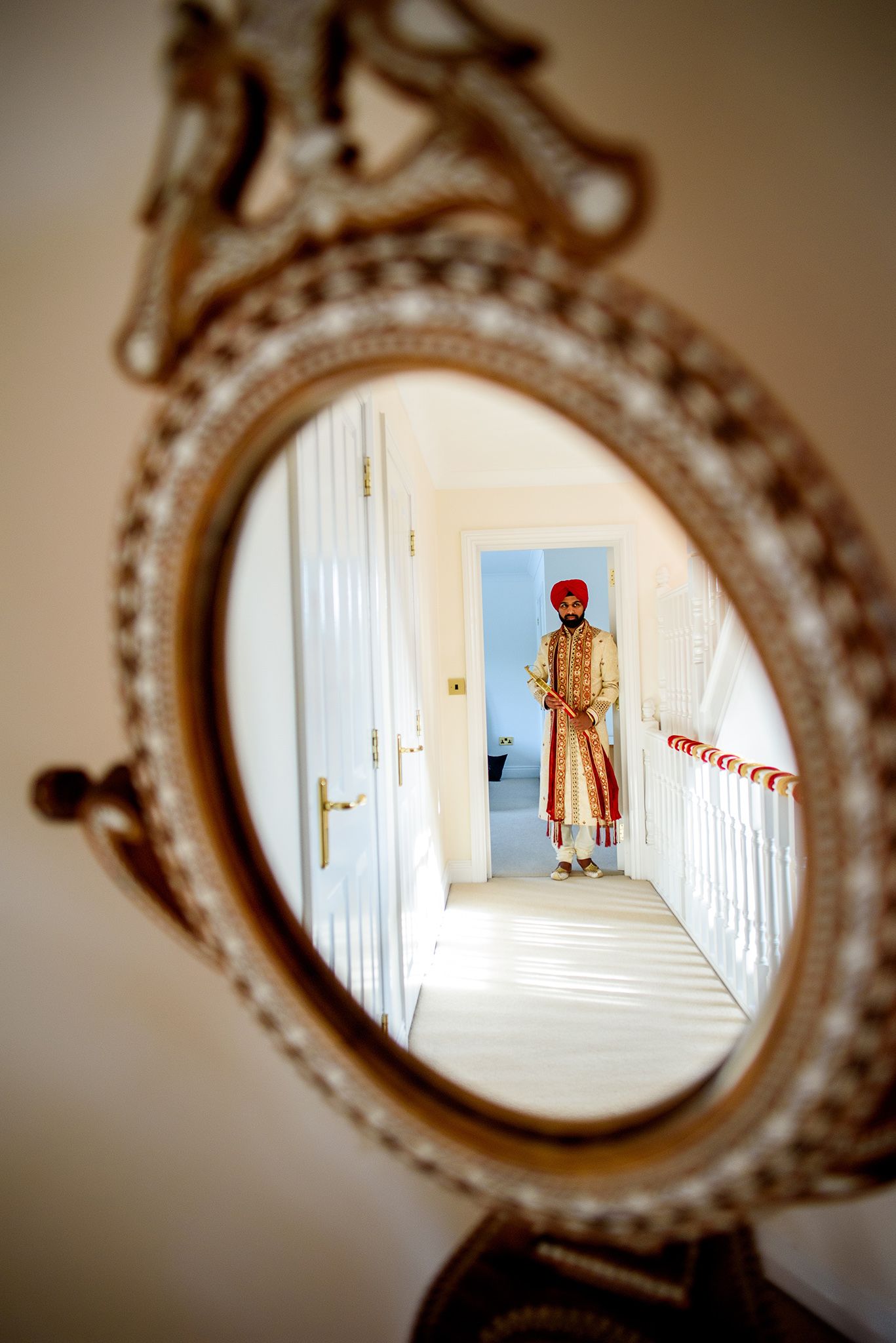 Ornate vintage mirror reflecting a traditional Indian groom in formal attire standing in a hallway.